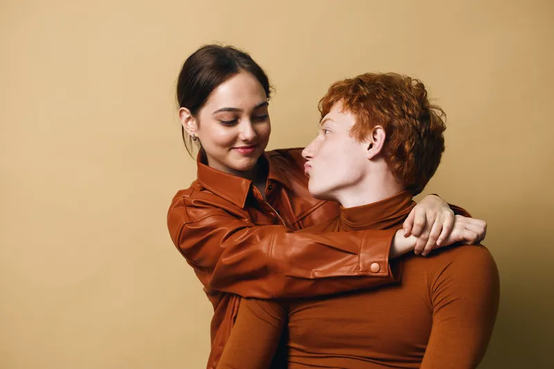 Couple embracing and smiling with affection against beige background. Young man and woman in brown clothing share tender moment and eye contact together.
