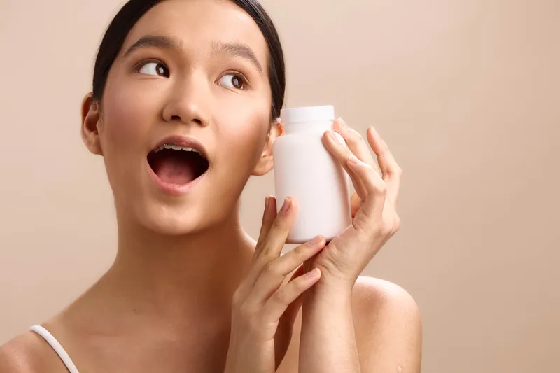 asian woman holds a white bottle up to her mouth with a bright smile in a soft warm setting, presenting a playful, healthy lifestyle moment that feels fresh.