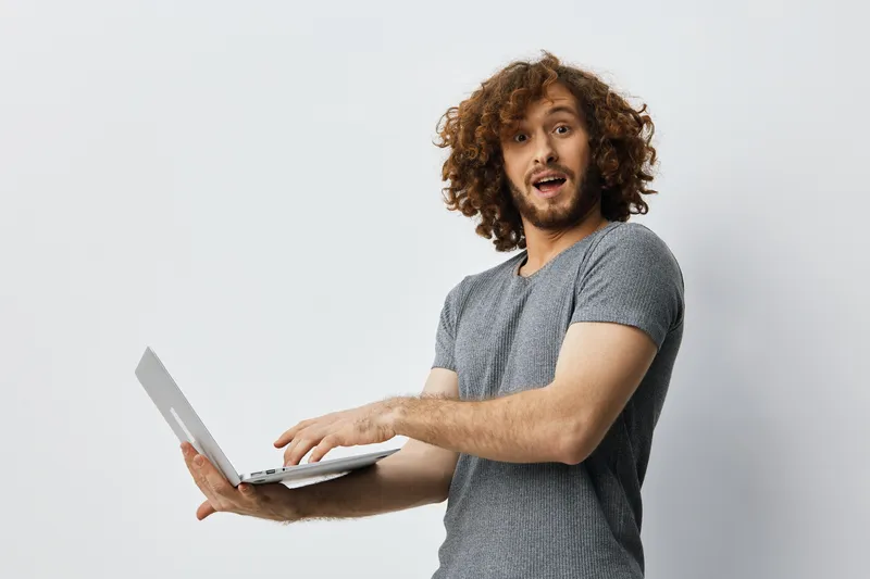 Excited young man with curly hair using a laptop and smiling in a bright, minimalistic setting, conveying enthusiasm for technology and communication