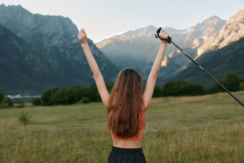 A cheerful person stands in a wide open field with arms raised, enjoying the outdoors. The distant mountains and clear sky create a hopeful, energetic atmosphere.