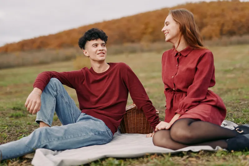 Candid young couple enjoying a relaxed outdoor picnic on a blanket with a wicker basket, exchanging genuine smiles and natural gestures in an authentic, credible autumn meadow scene.