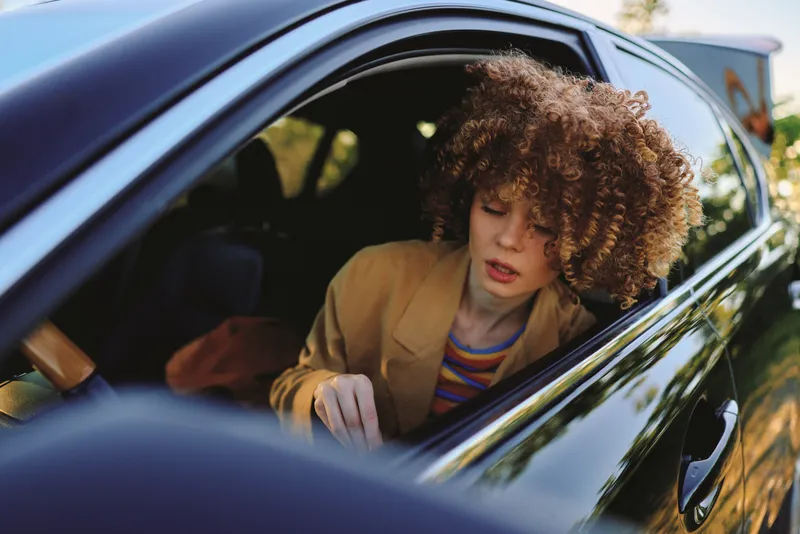 A woman with curly hair sits in a car, head tilted down, wearing a tan blazer. Retro film color tones create a nostalgic lifestyle moment and reflective mood.