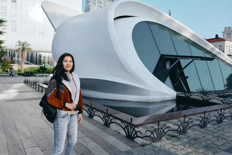 Woman beside a futuristic building with curved architecture in an urban outdoor setting, capturing travel lifestyle and fashion amid daylight and a striking cityscape scene.