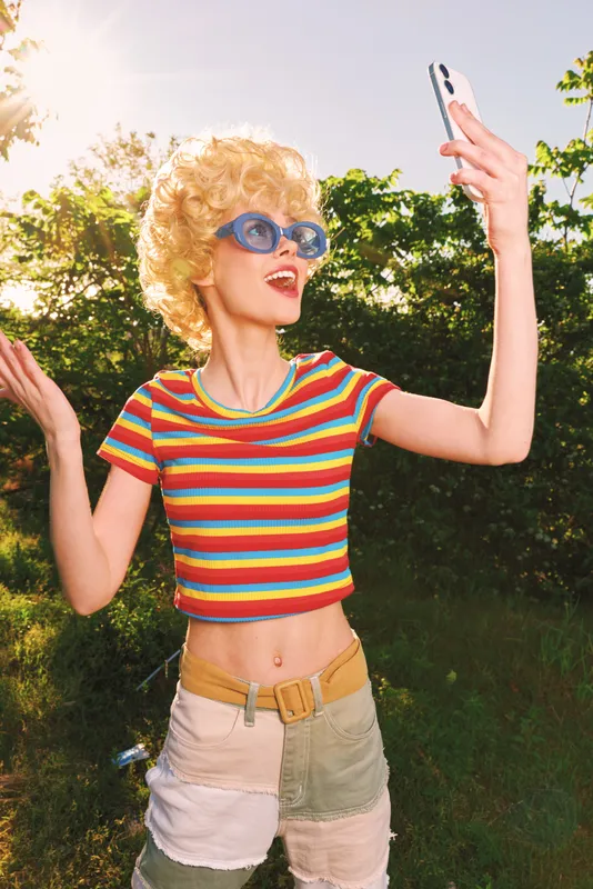 Young woman outdoors taking a selfie, retro film color vibe, striped crop top and denim shorts, bright sunglasses, sunny park setting, casual lifestyle moment captured with vibrant tones.