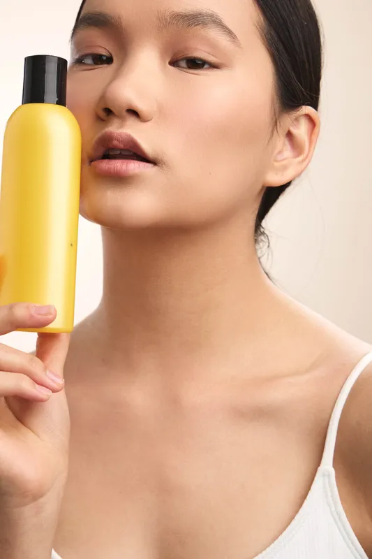 asian woman holds a yellow bottle close to her face in a closeup portrait, presenting a calm, confident vibe with clean styling and modern aesthetic.