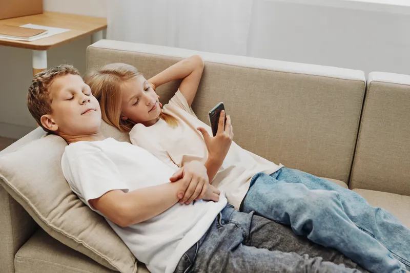 Two relaxed children lying on a couch in casual clothes, one holding a smartphone, enjoying leisure time indoors in a bright home environment.