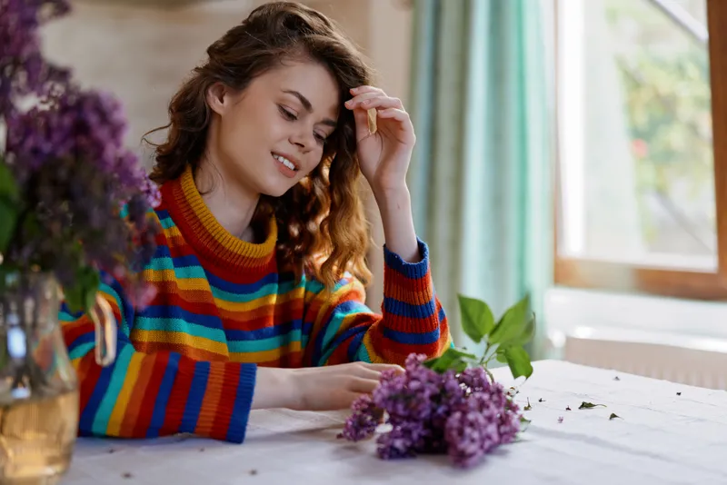 Young woman in a vibrant striped sweater sits at a sunlit table arranging lilac flowers, smiling softly in a cozy home setting