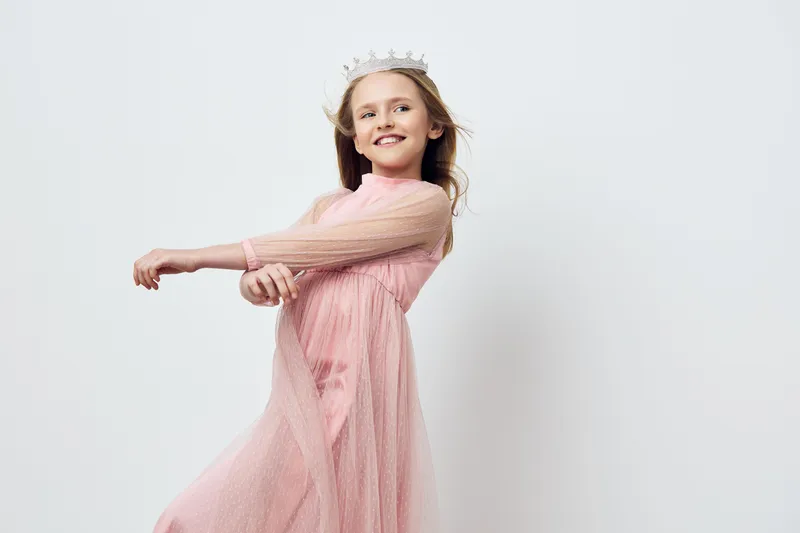 A cheerful little girl wearing a pink dress and crown, showcasing joy and playfulness against a minimalist white background Perfect for children s fashion and celebration concepts
