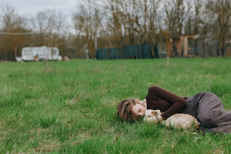 Relaxed woman lying on grass outdoors with a playful goat kid resting beside her, soft breeze, natural colors, serene countryside mood, candid lifestyle moment, warm sunlight, gentle companionship