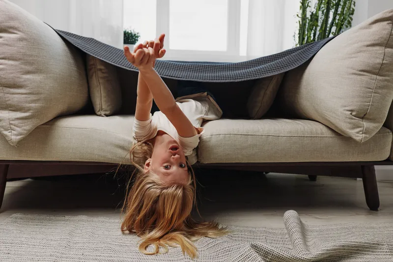 Playful child with long hair lies upside down under blanket on sofa in bright living room. Concept of childhood fun, home relaxation, and carefree moments indoors with natural light.