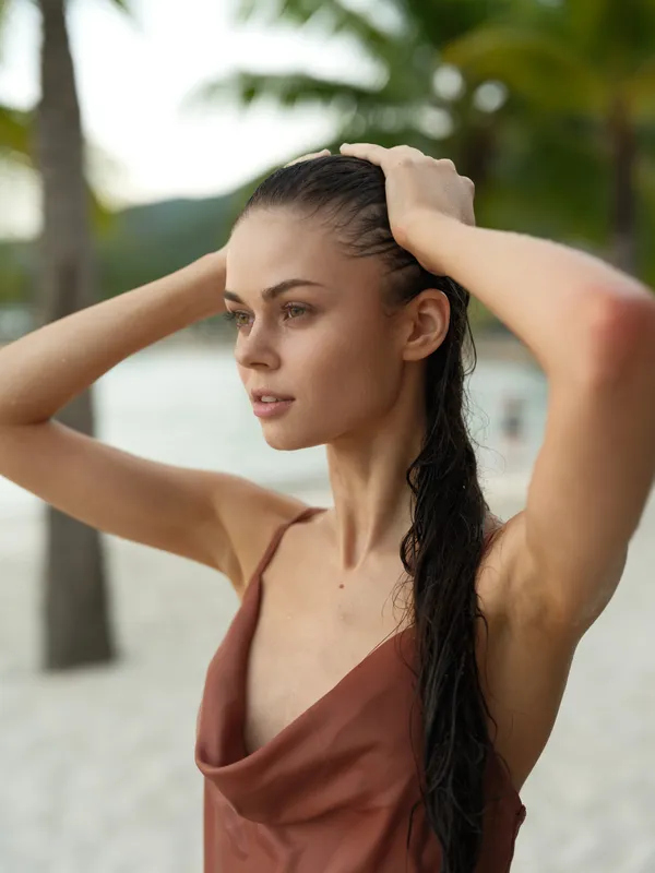 Young woman with wet hair on tropical beach, wearing a brown dress, looking thoughtful and serene, palm trees and soft sand background.