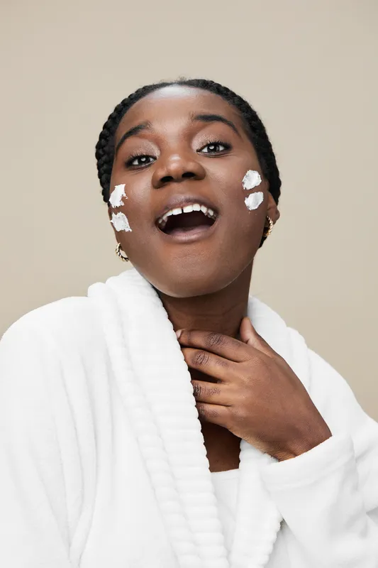 Joyful young woman applying skincare with cream on her face, showcasing beauty and self care in a minimalistic setting