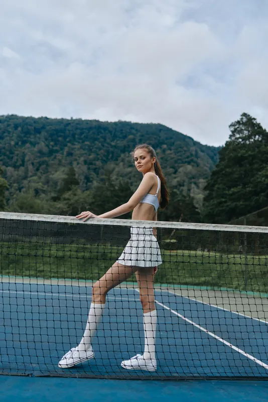 Athletic young woman in sportswear posing at a tennis court, showcasing confidence and style, surrounded by lush greenery in a vibrant outdoor setting