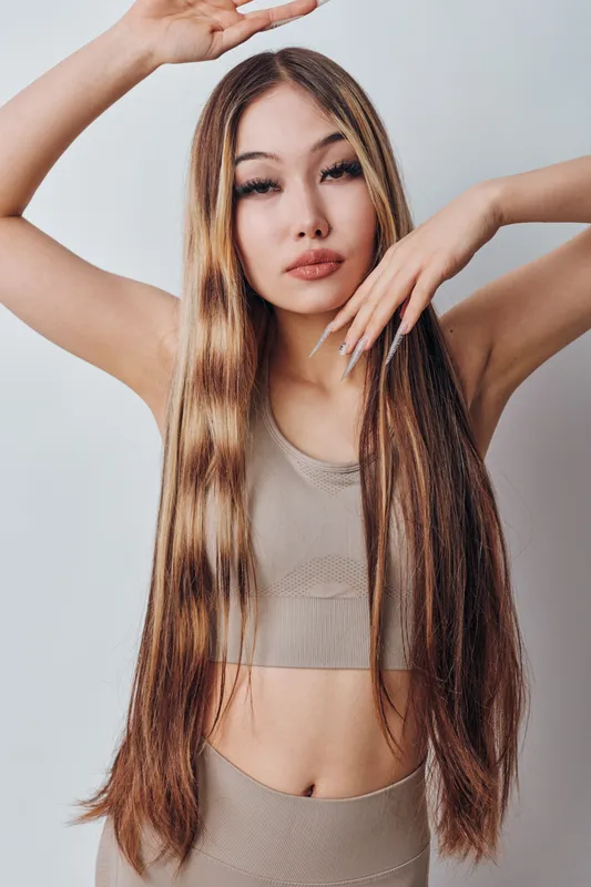 Woman with long straight hair posing with hands near face and head, wearing beige sleeveless top looking at camera on plain background in studio portrait fashion and beauty.