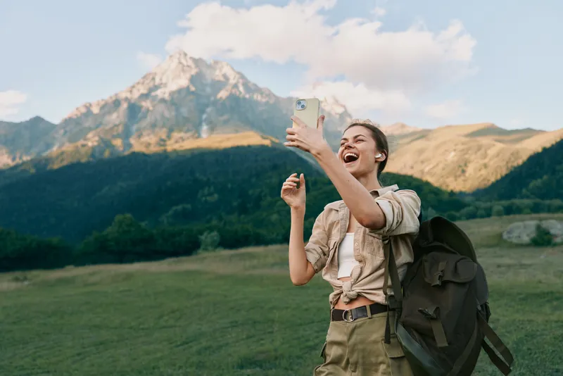 Happy woman outdoors taking selfie with smartphone, casual outfit, backpack, mountain landscape background, sunny day, natural light, joyful expression, travel concept