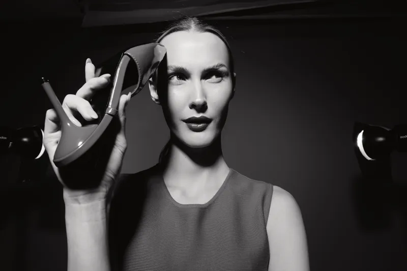 White woman with pale skin holds a high heel shoe near her face, posing confidently in a studio with dramatic black and white lighting. Concept of fashion and style.