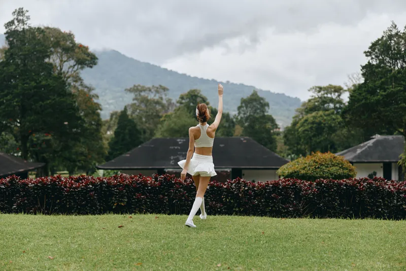 Cheerful young woman in white athletic wear dancing outdoors in a green garden, showcasing joy and fitness against a scenic backdrop of mountains
