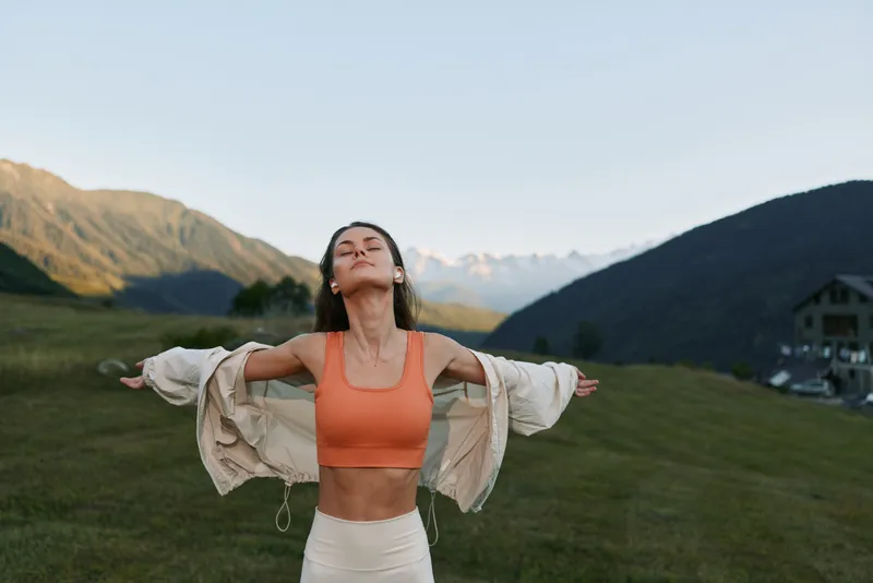 A joyful woman stands in a scenic meadow with mountains in the background, arms outstretched, eyes closed, enjoying fresh air and a moment of personal freedom.