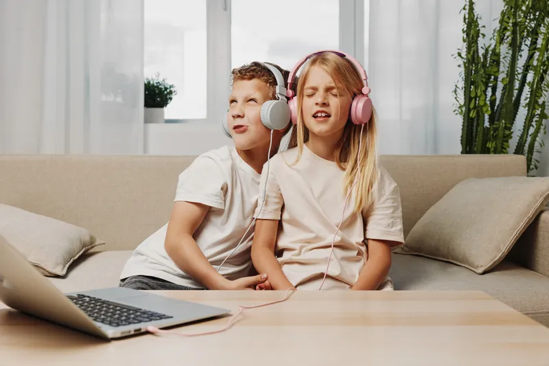 Two children wearing headphones listen to music together on a sofa in a bright living room, enjoying a shared moment of relaxation and fun.