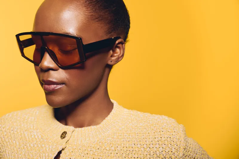 Stylish African American woman with dark skin wearing large angular sunglasses and a yellow knitted sweater against a bright yellow background. Concept of fashion and modern style.
