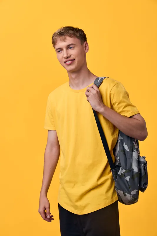 Casual young man in a bright yellow t shirt with a camo backpack over one shoulder stands against a vibrant backdrop, ready for school or travel, showcasing everyday style and confident energy.