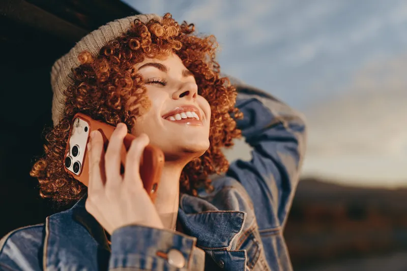 Curly hair woman smiling while holding smartphone outdoors in denim jacket, sunny lifestyle moment, joyful communication on phone with friends, soft sky background, candid close portrait