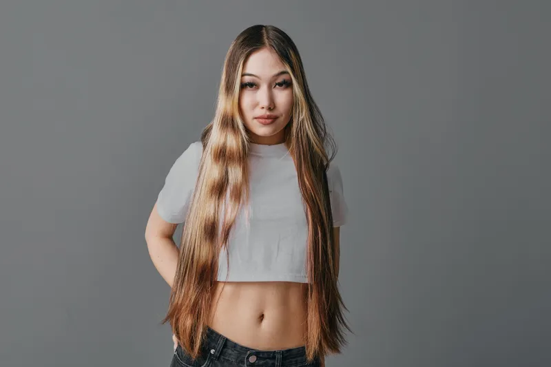 Young woman with long hair wearing white crop top and denim jeans standing against gray background, posing confidently with hands behind back.