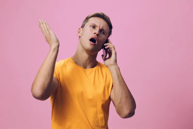 Frustrated man, middle-aged, with short light brown hair, wearing orange t-shirt, looking confused and annoyed while talking on smartphone, expression of disagreement, standing against pink background