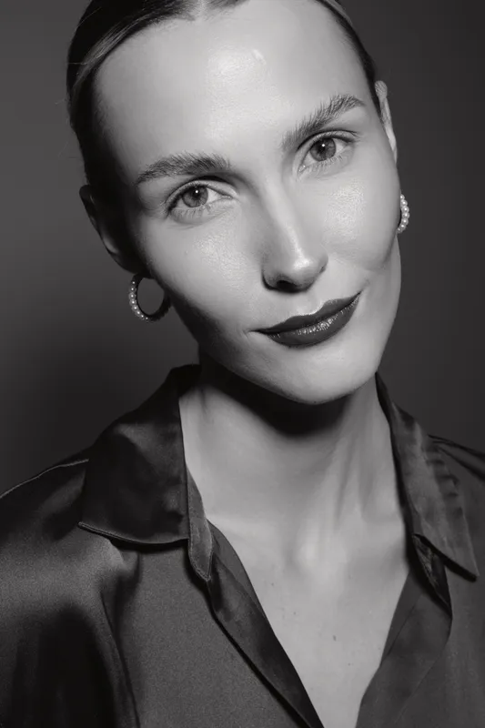 Close-up portrait of a Caucasian woman with fair skin and slicked back hair wearing hoop earrings and a satin blouse in studio lighting.