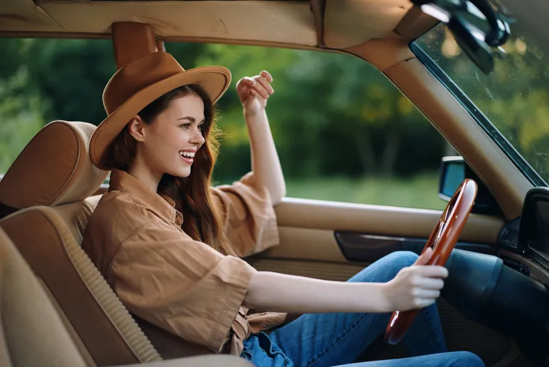 Young woman driving a vintage car with a joyful expression, wearing a brown hat and casual clothing, surrounded by greenery in a vibrant atmosphere.