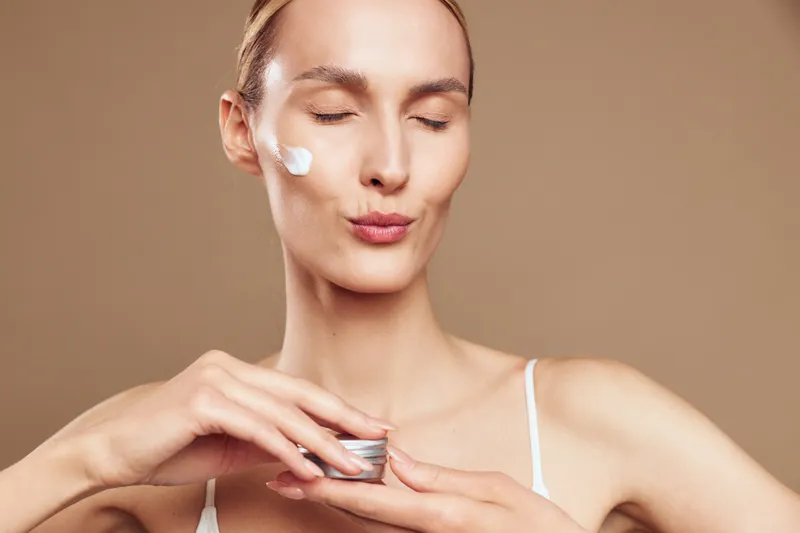 Young woman applies face cream on cheek with closed eyes enjoys skincare routine holds jar of moisturizer wears white tank top isolated on beige background.