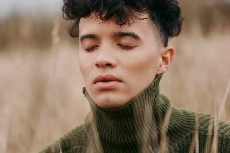 Close portrait of a young man in a green turtleneck standing in tall grasses with closed eyes, conveying quiet introspection and natural authenticity through soft light and textured skin.