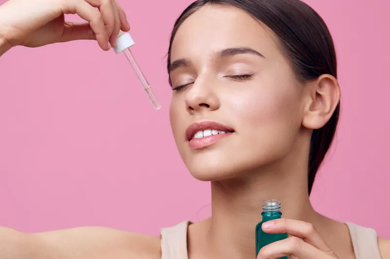 Young woman applying facial serum with a dropper on a pink background, showcasing skincare routine and healthy skin for beauty and wellness concept