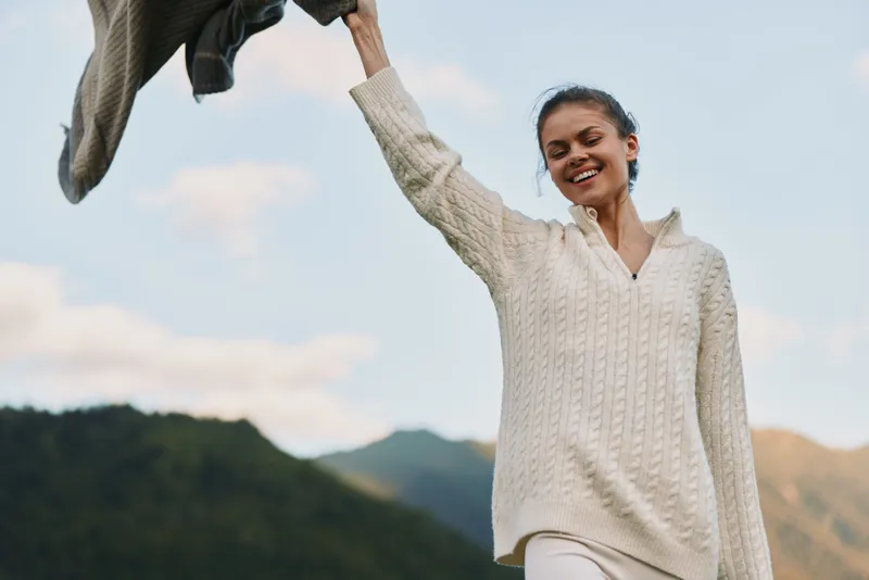 Young woman outdoors lifts a sweater in the air with a bright smile, enjoying a carefree moment in a mountain landscape