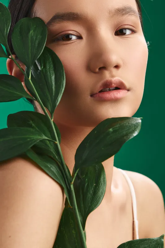 Asian woman gazes softly at the camera with lush green leaves near her face, creating a fresh, natural portrait in a calm studio setting that emphasizes harmony and serenity.