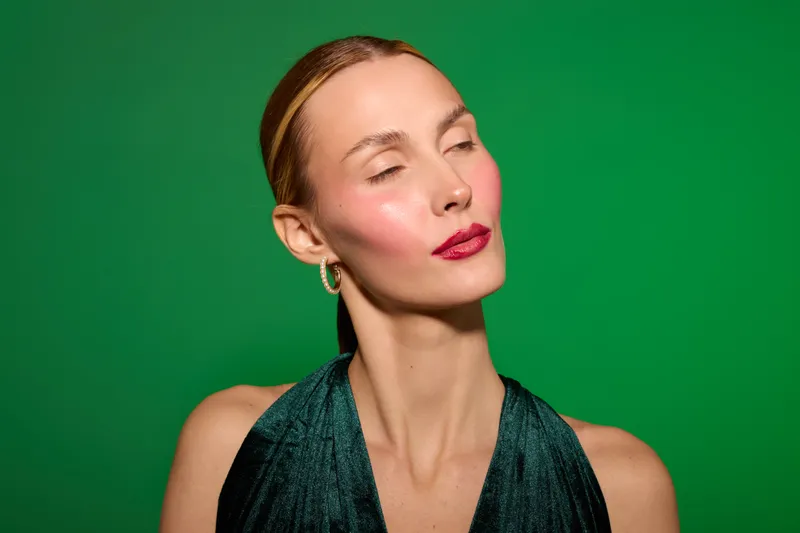 Portrait of a confident woman with closed eyes wearing a dark green dress and gold earrings against a green background. Concept of elegance, beauty, and calmness in fashion.