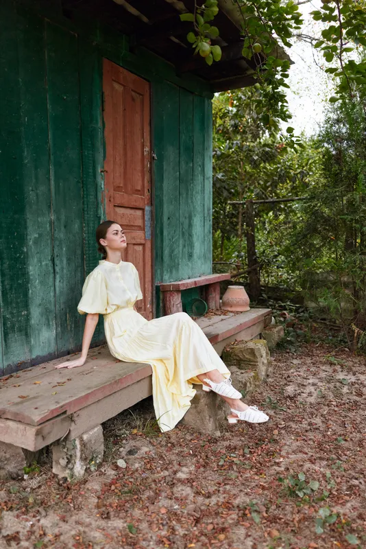 Woman in a yellow dress sits on a wooden bench beside a green door in an outdoor garden setting, exuding calm and relaxed fashion vibes, surrounded by natural light and tranquil scenery.