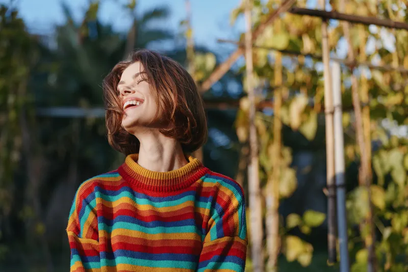 Young woman smiling in a colorful striped sweater outdoors in a tropical garden, short brown hair laughing while looking up, casual outfit and sunlight lifestyle portrait for travel and joy