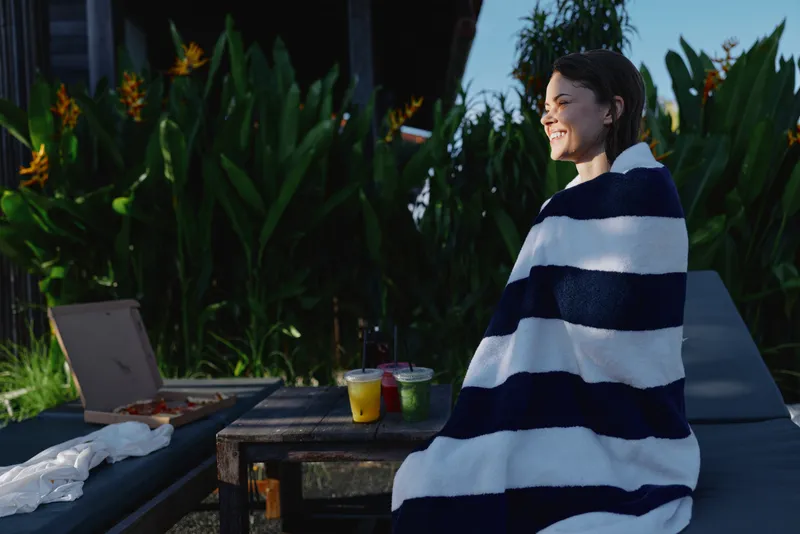 Poolside scene with a young woman wrapped in a striped towel, working on a laptop while pizza and drinks sit nearby, a relaxed sunlit lounge moment