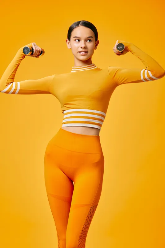 Female fitness model in bright orange athletic wear lifts dumbbells in a studio with a bold orange backdrop, showcasing strength, balance and a dynamic workout routine.