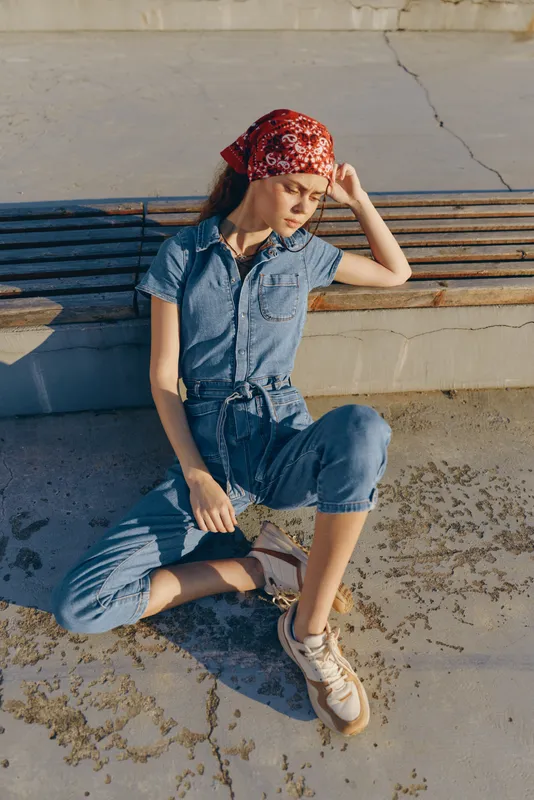 Lifestyle woman in modern boho-western denim street style with a red bandana sits thoughtfully on urban pavement, film color tones enhancing the relaxed atmosphere.