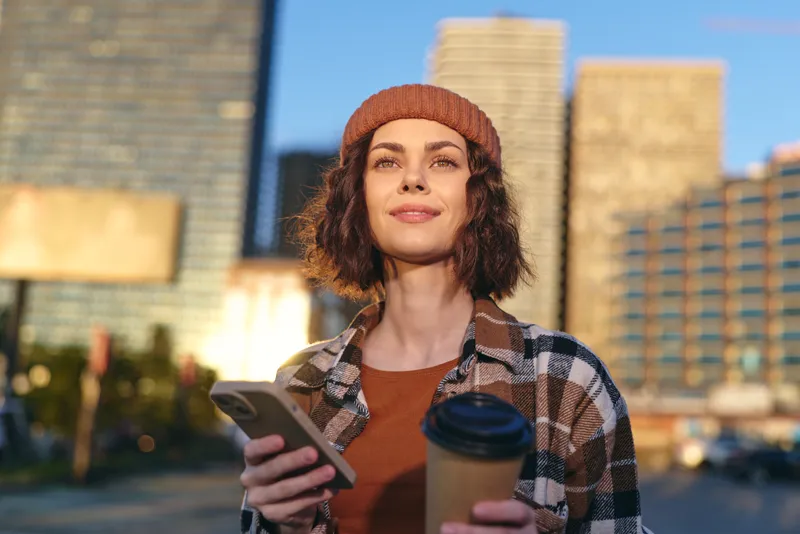 woman with coffee and smartphone in city portrait, smile and beanie in warm sunlight, authenticity and candid lifestyle, golden hour glow, mindful living and emotional storytelling urban scene