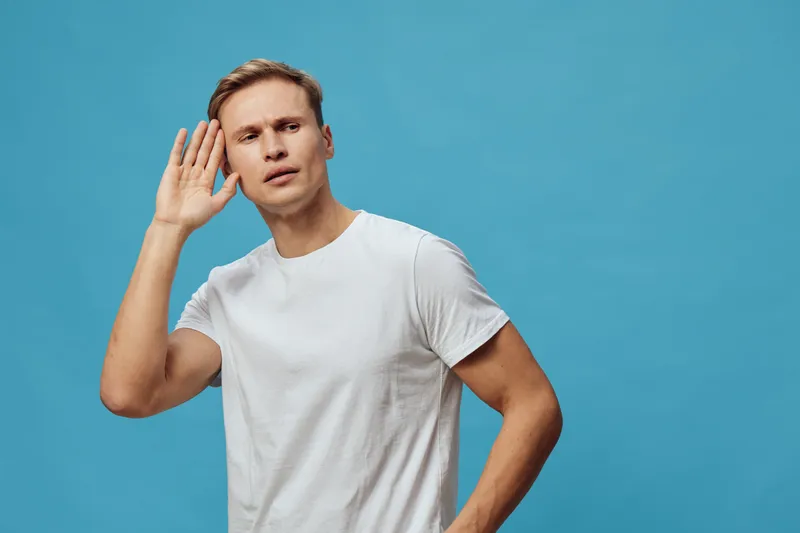 Young caucasian man wearing white t-shirt listen hand near ear looking curious isolated blue background concept mental health and gadgets