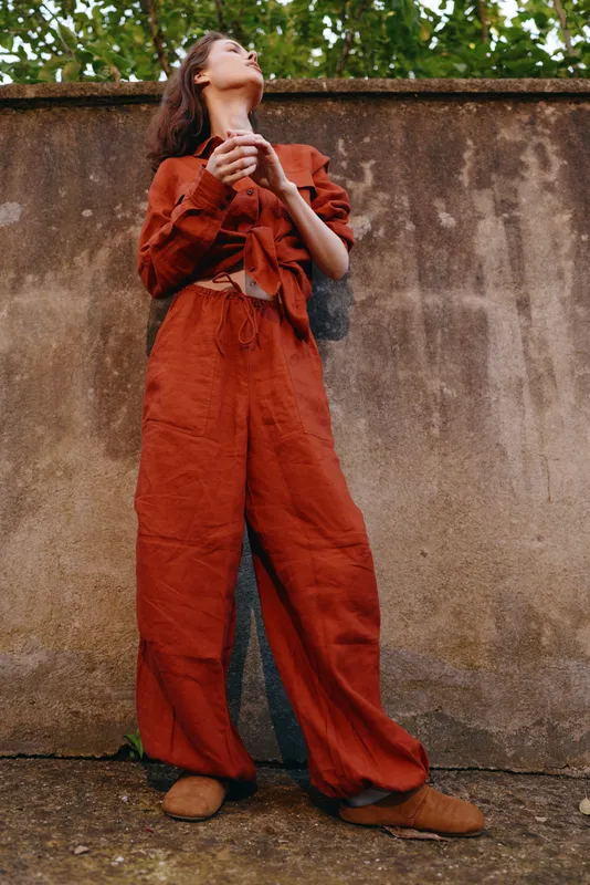 Casual woman outdoors wearing a bold orange jumpsuit, standing against a weathered wall, looking upwards with a serene expression, natural light, relaxed vibe, lifestyle focus