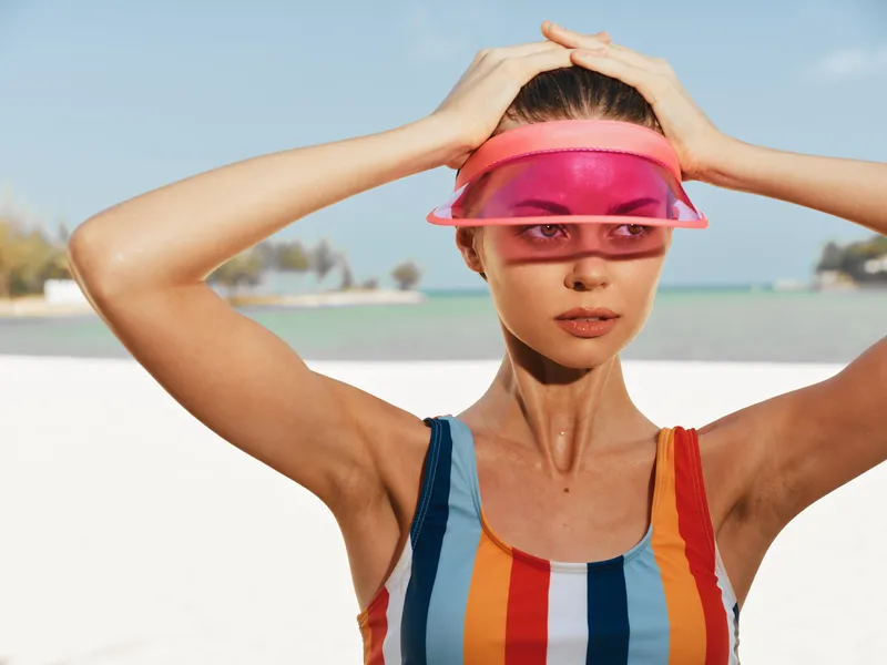 Portrait of a woman on a sunlit beach, adjusting a vivid pink visor over her eyes, wearing a colorful striped swimsuit. The scene exudes bold summer style and sunlit energy, with a soft breeze, clear