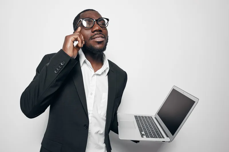 African American man with dark skin wearing glasses and black suit talking on phone while holding laptop in white studio. Business communication and technology concept.
