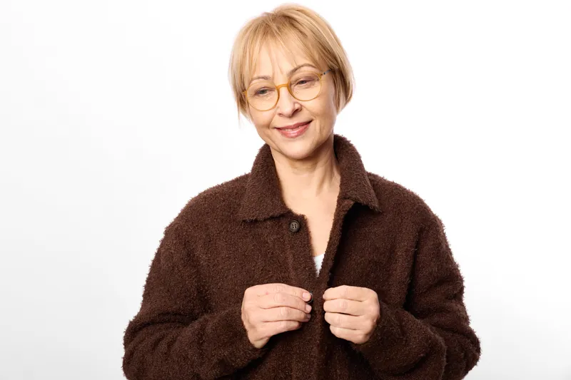 Woman, elderly, glasses, smile, portrait, coat, winter, studio background, short blonde hair and round glasses, dark brown wool coat, hands adjusting collar, warm calm mood, soft light