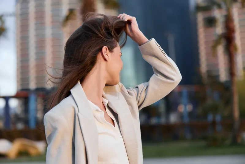 Woman in beige blazer touching hair outdoors with blurred urban background, natural light, casual confident pose, autumn fashion style