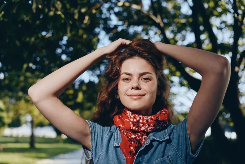 lifestyle woman in modern boho-western denim street style with curly hair and red bandana, natural sunlight, film color, outdoor park setting, relaxed mood.