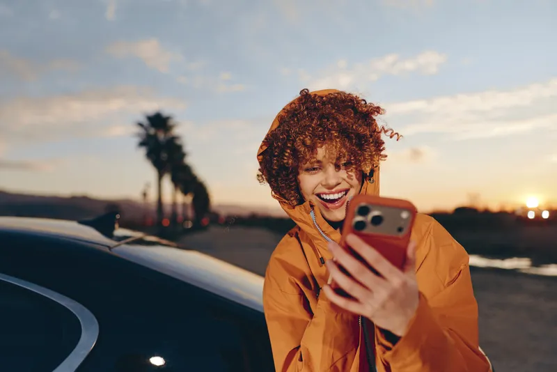 Woman lifestyle outdoors, wide smiling selfie smartphone in hand, curly hair in breeze, warm sunset light, orange jacket, parked car nearby, relaxed joyful mood, evening street background